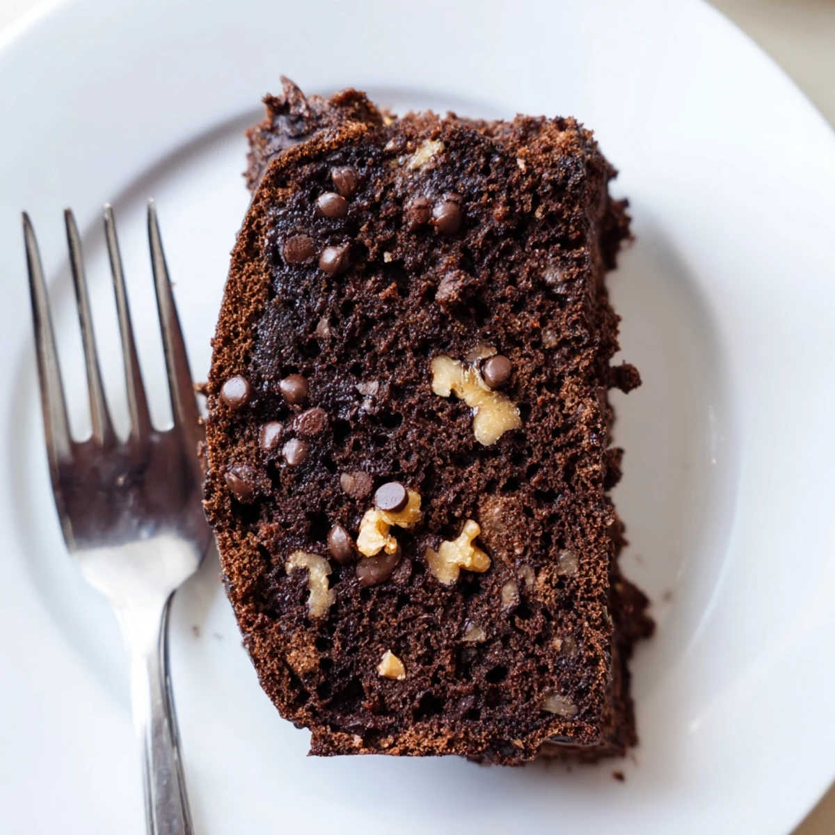 A thick slice of moist Chocolate Banana Bread on a white plate, with a dollop of whipped cream and a hot mug of coffee nearby.