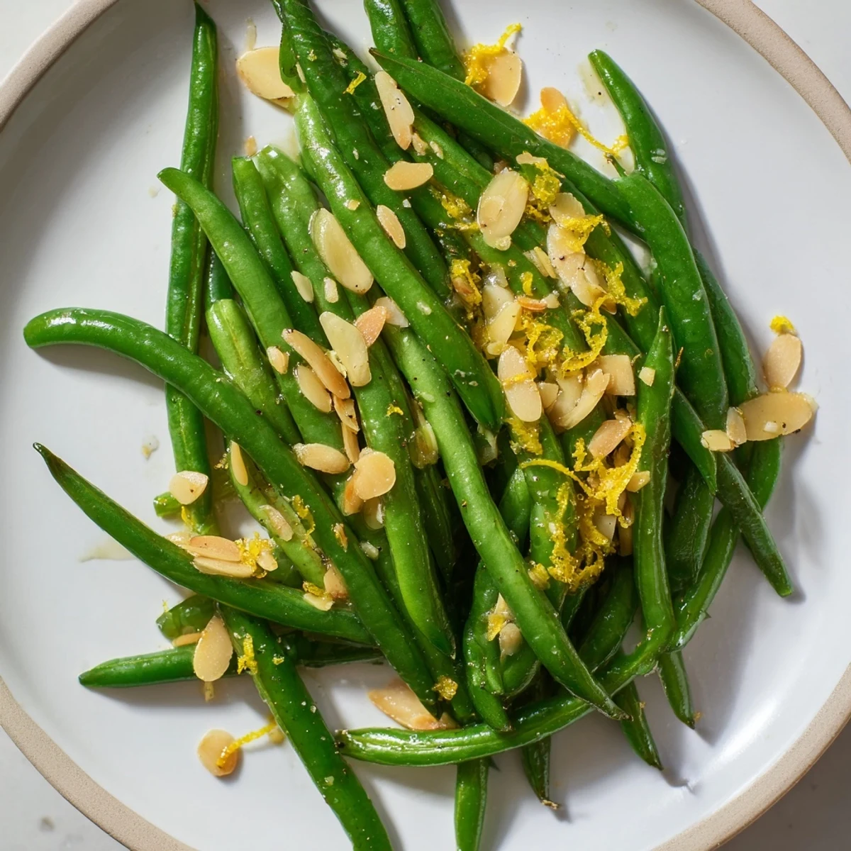 Tender green beans with lemon and almonds served warm on a white plate garnished with zest.