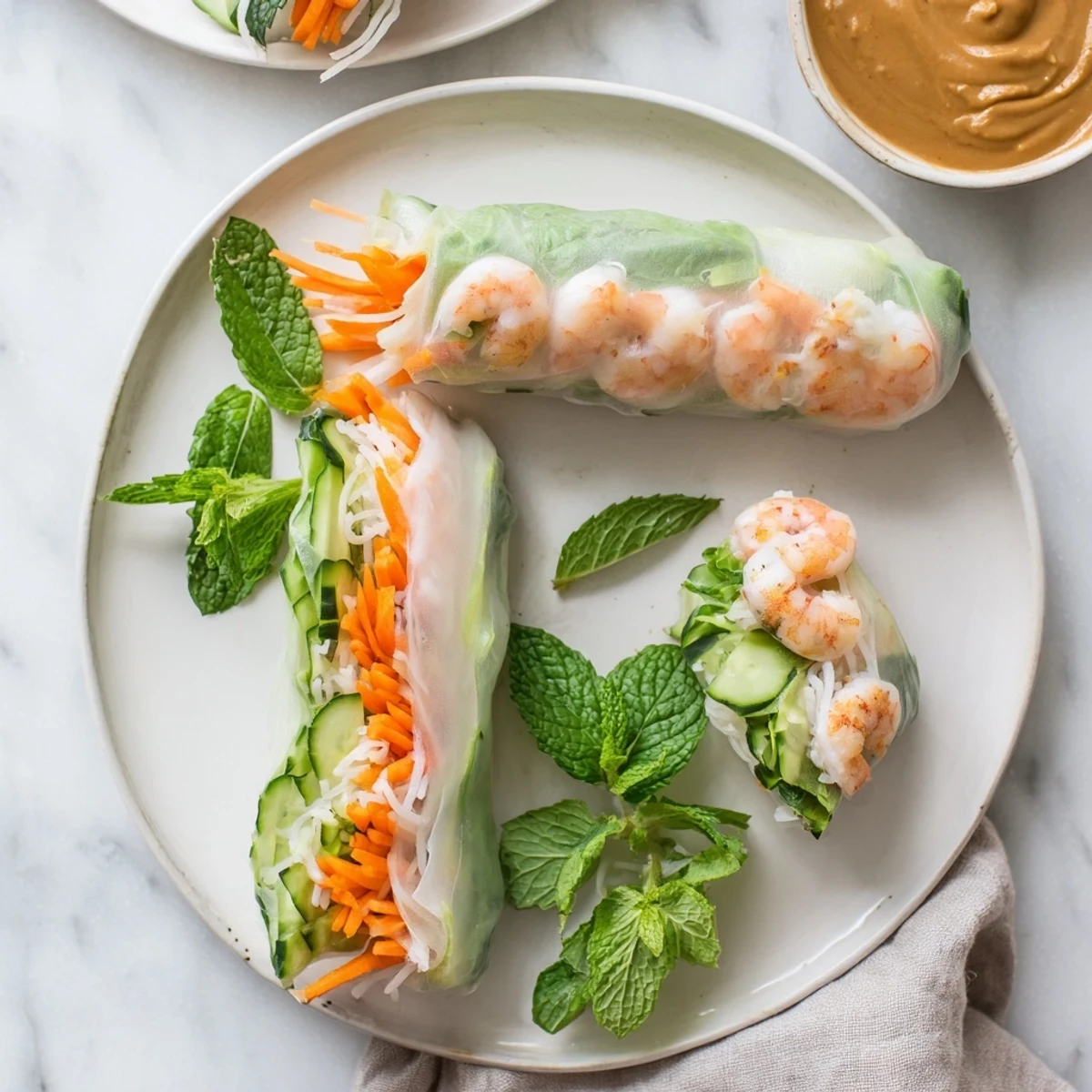 Freshly rolled Vietnamese Spring Rolls with crisp carrots, cucumbers, and succulent shrimp, served with peanut dipping sauce on a white plate.  