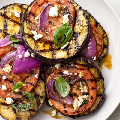 Charred grilled tomato eggplant layered with red onion and drizzled with balsamic marinade on a rustic plate